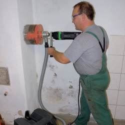 mann in grünem overall bohrt mit eibenstock kernbohrgerät mit staubsaugeraufsatz loch in weiße wand
