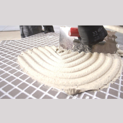 hand mit schutzhandschuh zieht hellgrauen fliesenkleber mit gezahnter kelle auf kariertes fliesenrückwandpaneel